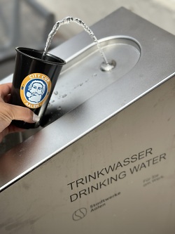 Auf dem Bild ist ein öffentlicher Trinkwasserbrunnen mit dem Aalener Mehrwegbecher City Cup mit Spion Motiv zu sehen.